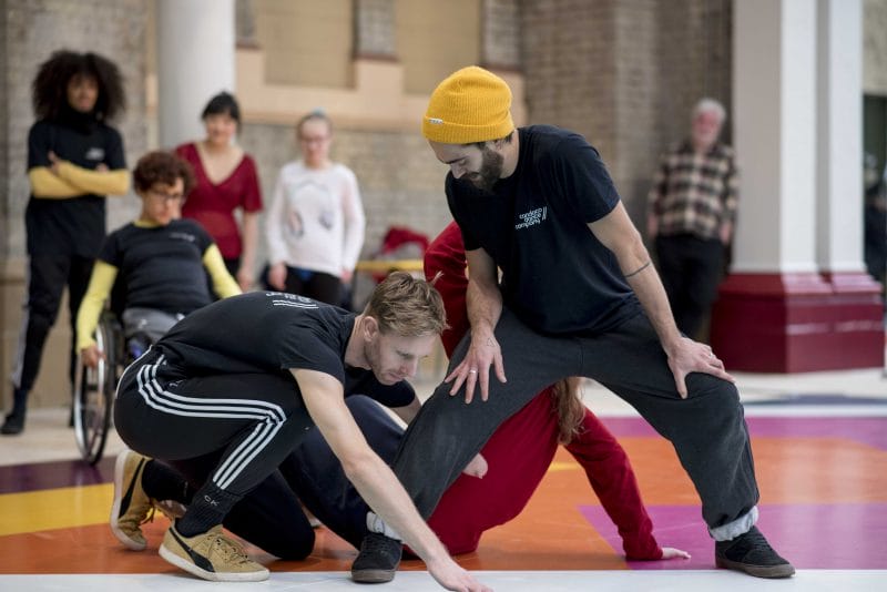 2 dancers lean into each other, one standing the other crouching