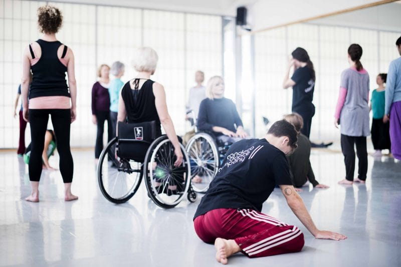Candoco dancer kneels with head down in the foreground, dancers using wheelchairs and others standing in the background