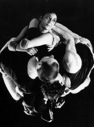 black and white image of dancer making a circular shape as they wrap their hands around each other, one dancer looks up, the others have their heads down