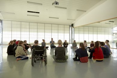a large group of people sit in a circle in a light white studio.