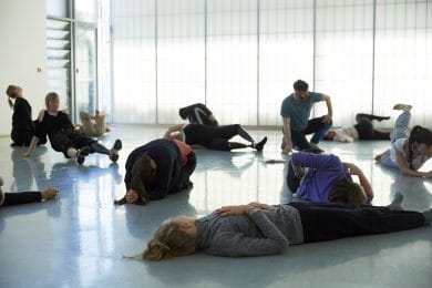 Several dancers lie on the floor in various positions in a white, light studio.