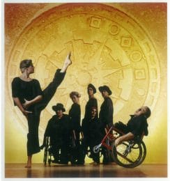 A group of disabled and non-disabled dancers stand on stage in front of a yellow background. A female dancer stands facing a male dancer in a wheelchair with her left leg lifted and pointed