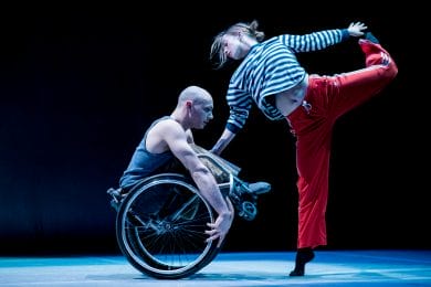 A man is sitting in a wheelchair. A woman is holding the left wheel whilst holding her leg up with her left.