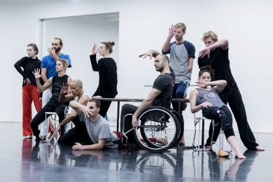 a group on disabled and non- disabled dancers stand in a group with a table in the middle. They all stare at one point ahead.