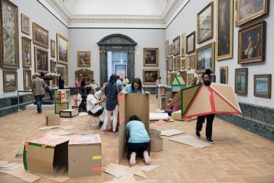 Groups of people scattered in an art gallery room building with carboard and tape.
