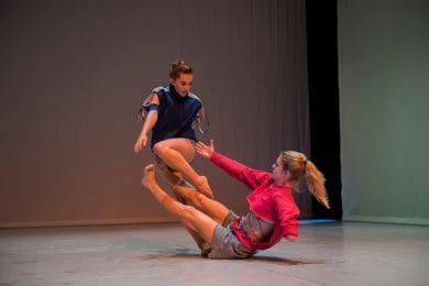 2 dancers are on a grey stage. One is on the floor with their legs stretched out in front of them and their right arm reaching to another dancer. This dancer is staring at the hand and has their left leg bent and their body twisted towards the person on the floor.