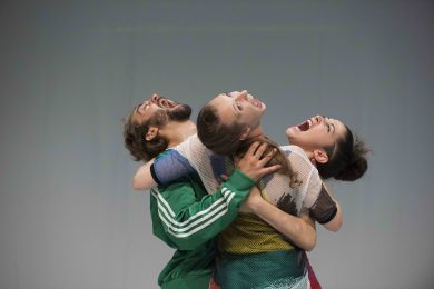 3 people in brightly coloured clothes embrace. Their heads are lifted up with their mouths wide open.