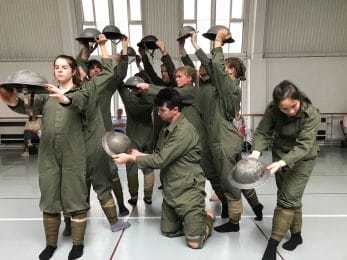 Group of dancers dressed as soldiers in green boiler suits, stretch arms out and up holding a metal army helmet