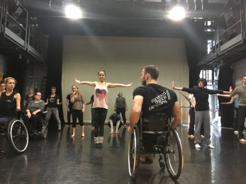 a group of dancers stand in a studio with their arms stretched out. A male dancer sits in a wheelchair with his back to the camera, looking at the group.