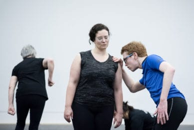 In the forefront, two dancers lean against each other. A third dancer in the background raises one elbow in a white walled studio.