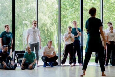 A led Artist teaching a to a group of young participants in a bright studio space. The photo is captured from the teacher's back and the students are standing in front of a large glass window.