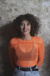 Headshot of Anna Seymour, smiling with short curly brown hair. They are wearing an orange top with a black bra visible underneath. Anna describes herself as follows: Anna has dark brown curly hair just above her shoulders with a fringe, brown eyes, fair skin with freckles, a nose ring and has a petite frame. Anna self-describes as: a 40 year old white woman with dark curly hair that comes to just above the shoulder and a wavy fringe. They have a nose ring an freckles. They are D/deaf and have a petit frame.