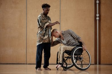 Two male dancers. One stands to the left of the image with his front elbow raised and lower arm across his body. This arm is above the head of the second dancer who is a wheelchair user. His head leans against the standing dancer's torso.