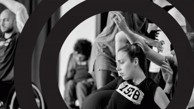 Left - dancer using wheelchair raises arms, front - dancer is on the floor gazing to the left with elbow bent up and hand resting on an arm that is entering the shot from the right. Other bodies are moving in the background.