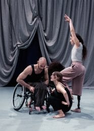 3 people in front of a grey draped curtain which is hoisted up in the centre. Liv is in the front and is crouching down, Joel is seated in a wheelchair and leaning forwards towards Liv. Mickaella is standing to the side with her front knee lifted and arms long above her ahead.