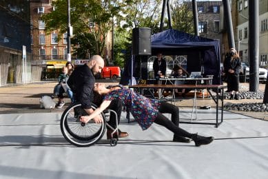 Day of Dance. Two Candoco dancers perform outside. Joel has a beard and a shaved head. He catches Anna in his wheel chair. Anna has dark curly hair and is falling backwards into Joel's lap