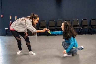 Taster session. Two dancers face each other. One has brown hair in a pony tail and is bending towards the other. The second dancer is kneeling on the floor. They each have one arm stretched towards the other and their hands are touching. They are in a dance studio with a light grey floor and dark blue walls