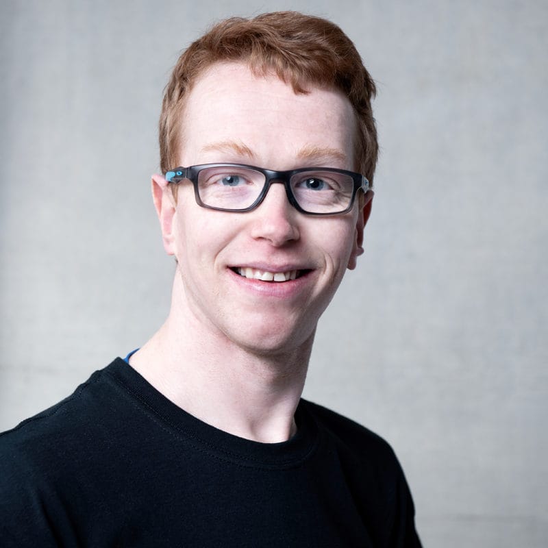 Dermot Farrell. A smiling headshot of a dancer with short red hair and black framed glasses. He is wearing a black tshirt.