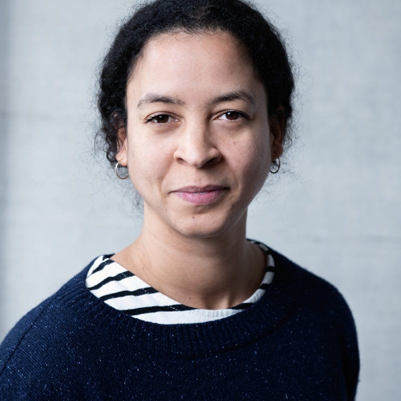 Vanessa Abreu. A smiling woman with dark hair is wearing a dark blue jumper. We can just see a white tshirt with black strips appearing at the neckline