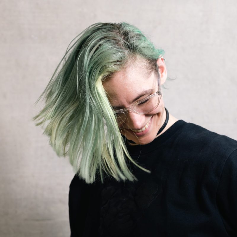 A person with light green hair and a black top is smiling and looking down.