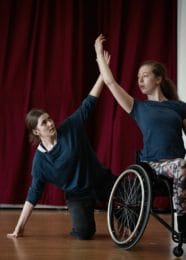 One dancer in a wheelchair looks to her left with her arm raised in the air, another dancer kneels on the floor behind her with her left hand supporting her and her right hand in the air almost touching. Both dancers in front of a dark red curtain.
