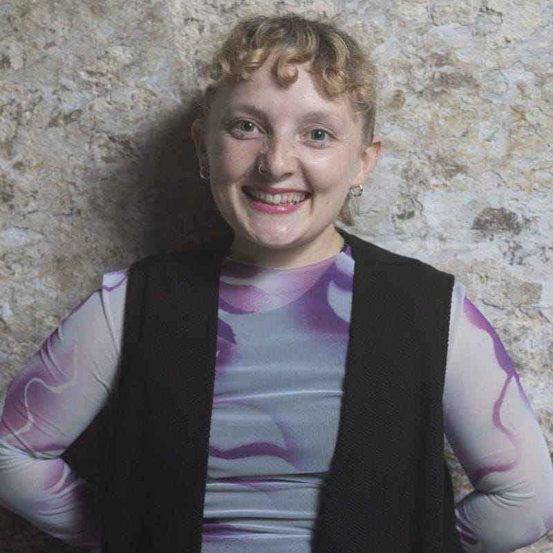 Headshot of Annie Edwards, a person with short, blonde curly hair, smiling at the camera. They are wearing a grey and purple top layered under a black gilet jacket, with their arms positioned behind their back. Annie self-describes as: a small white woman in her 20's with short curly hair.