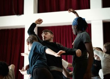 3 dancers working in a trio. One dancer is turning around with their left arm lifted in the air.