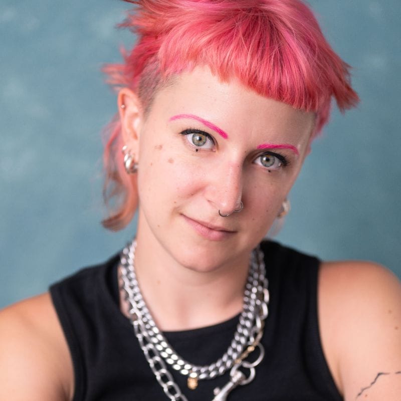 Jay is looking into the camera with a blue background behind them. They have short pink hair and they are wearing a black vest top with some silver necklaces.