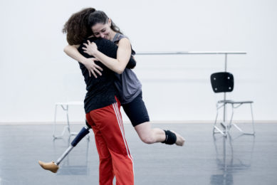 Two people hugging in a bright studio space. One person is lifting the other off the ground. The person who is being lifted is looking down smiling whilst facing the camera. In the background there is a ballet barre and two chairs.