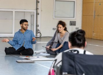 Photo taken behind someone's shoulder who they are looking at two people in front of them. The focus is on the two people who are sitting on the floor. One is looking down and holding a laptop in her hands whilst the other person is engaged in a conversation and moving their hands. On the floor there are paper sheets and note books.