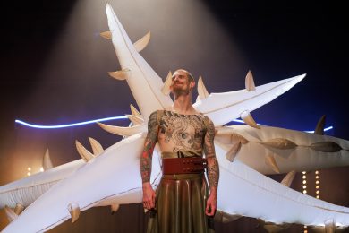 A person wearing a costume with large, white inflatable wings stands confidently, smiling and looking to the side. They are shirtless and have a large tattoo across their chest and arms, and they are wearing a dark gold skirt with a thick red belt around their waist.