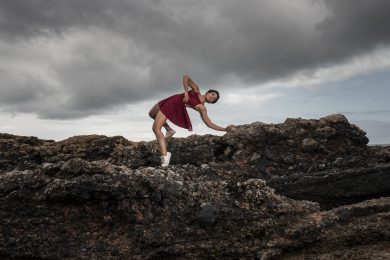 Dancer in a red dress and white shoes gracefully dances on rocky terrain, leaning elegantly to one side.