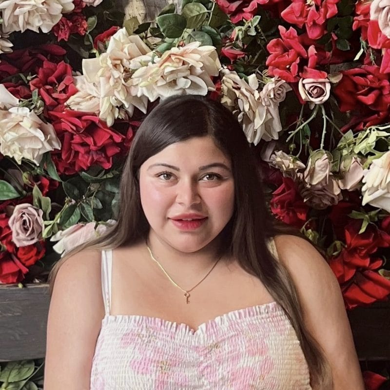 Headshot of a person with long brown hair and a pink dress looking straight at the camera, with red and white flowers in the background.