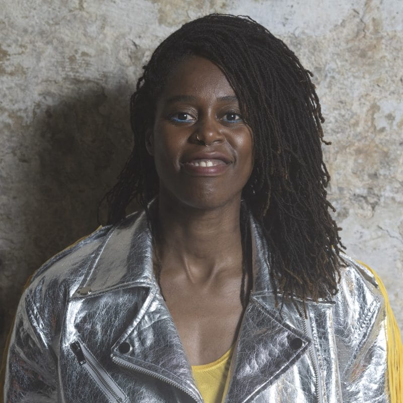 Headshot of Temitope Ajose, a person with mid-length dreadlocks, wearing a bright silver jacket over a yellow top, and smiling warmly at the camera. Temi self describes as: a tall black woman, in her mid-40s with mid-length, small sister locks.