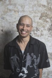 Headshot of James Olivo, a person with a shaved bald head and wearing earrings and a black long-sleeve shirt. They are smiling and looking slightly to the side, away from the camera. James self-describes as: a Filipino-Chinese person in their late 20's, with a shaved head.