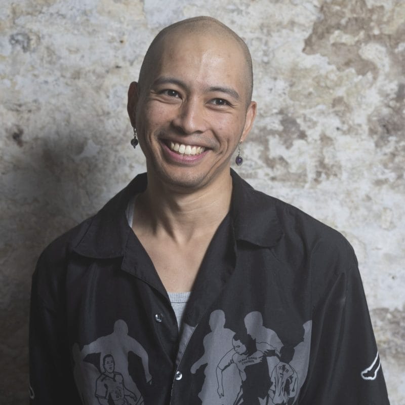 Headshot of James Olivo, a person with a shaved bald head and wearing earrings and a black long-sleeve shirt. They are smiling and looking slightly to the side, away from the camera. James self-describes as: a Filipino-Chinese person in their late 20s, with a shaved head.