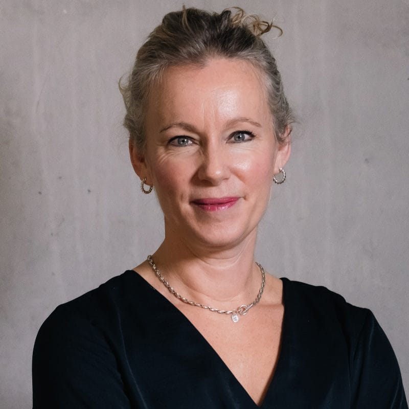 Headshot of a person with blonde hair neatly tied back, wearing a black v-neck shirt. They are looking directly at the camera with a gentle smile.