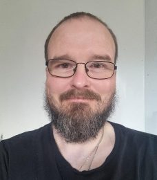 Headshot of person with glasses, short hair and beard, smiling at the camera. They are wearing a black t-shirt standing against a plain white wall.