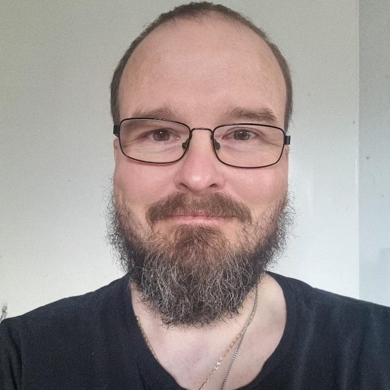 Headshot of person with glasses, short hair and beard, smiling at the camera. They are wearing a black t-shirt standing against a plain white wall.