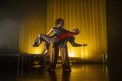 Two dancers perform on stage against a yellow curtain in the background. One dancer, with a strong and steady stance, lifts the other off the ground. The dancers look at each other with the standing dancer positioned in front of the camera.