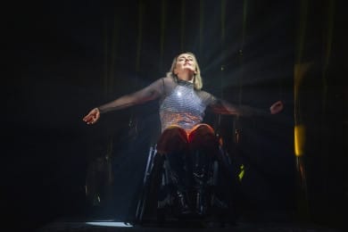 A performer dancing on stage with arms outstretched and eyes closed, radiating emotion. Their silver, sparkly top catches the light, creating a dazzling shimmer against the dark stage backdrop.