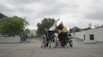 Two people in wheelchairs dancing next to one another outdoors. They lean on each other, one person facing the camera and one facing away from the camera.
