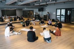A group of participants engaging in a workshop, all sat on wooden floors with large paper sheets scattered in the middle.