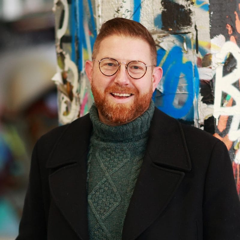 A person wearing a black jacket and high-neck jumper stands against a wall in a graffiti tunnel and smiles at the camera.