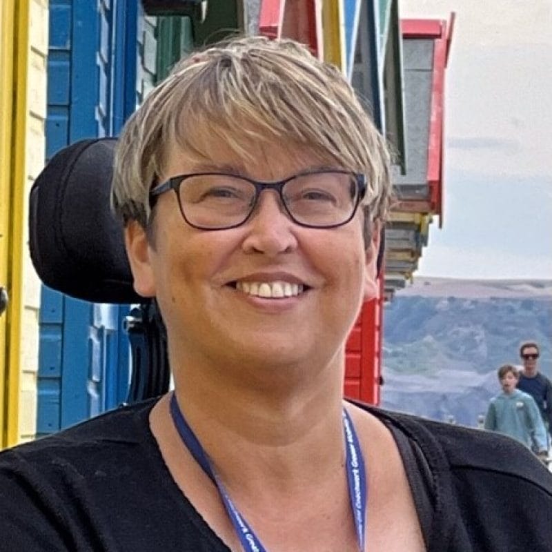 Headshot of person with short blonde hair, wearing a black shirt with a blue lanyard, smiles at the camera.