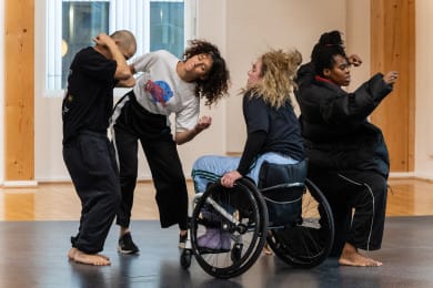 Four dancers move fluidly in a rehearsal studio, some with their eyes closed. Their bodies lean into one another, each facing a different direction, creating a dynamic and expressive composition.