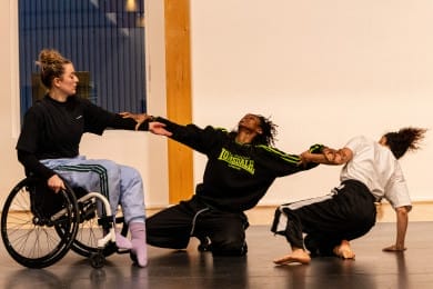 Three people dancing together. The central dancer leans back with arms outstretched, while the two dancers on either side hold their arms, pulling gently in opposite directions. Their expressions and movements convey rhythm and connection.