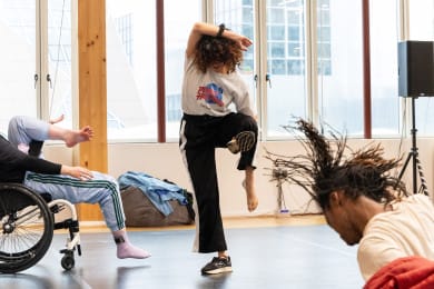 Three dancers are rehearsing in a studio, with the central dancer as the focal point. The dancer balances on one leg while holding their other leg up, with their shoe in one hand, while their other arm rests on their head.