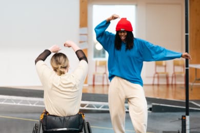Two people dance in a rehearsal studio - one seated turned away from the camera and one standing facing the camera. In the background, an L-shaped platform is positioned within the studio space, serving as stage design.