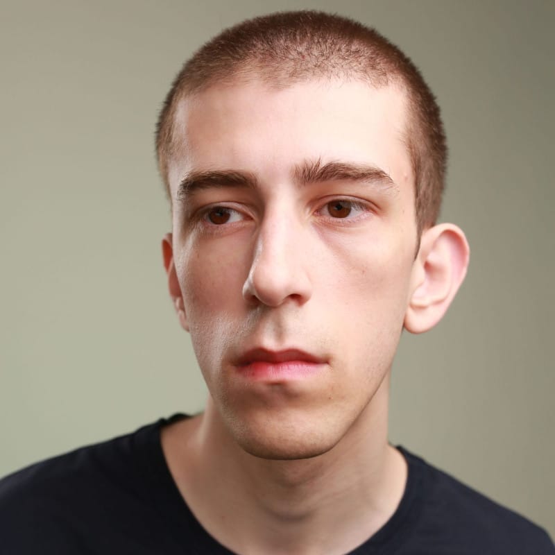 Headshot of a person with short hair, wearing a black shirt, looking away from the camera with a serious expression.
